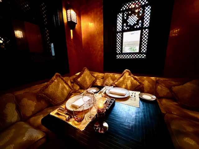 Elegant dining area of Le Marocain at La Mamounia, featuring traditional Moroccan decor with intricate wooden carvings, glowing lanterns, and an open courtyard surrounded by lush greenery.